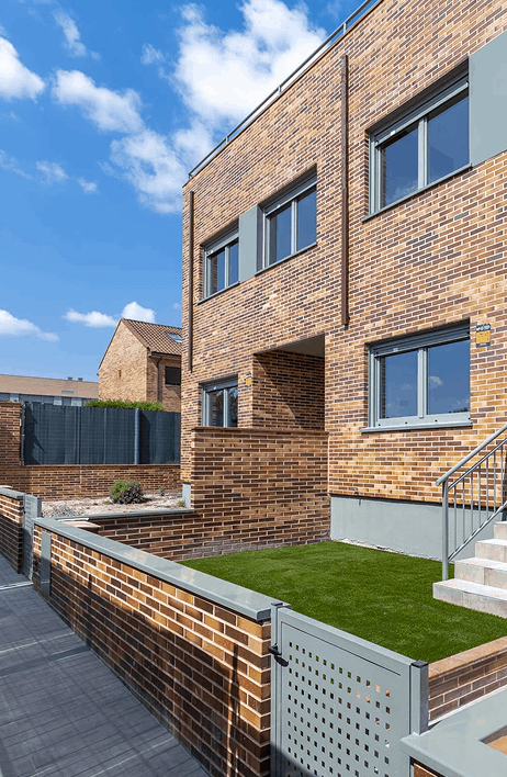 Vivienda adosada de obra nueva con fachada de ladrillo visto, ventanas modernas de aluminio y jardín privado con césped artificial. Urbanización residencial tranquila y bien cuidada, ideal para familias.