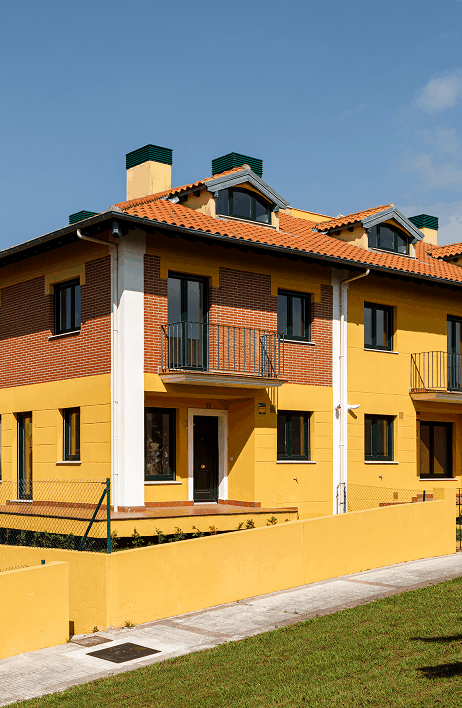 Vivienda adosada de dos plantas con fachada en tonos amarillo y ladrillo visto, tejado de teja roja y balcones con barandillas metálicas. Ubicada en una zona residencial con césped y acera peatonal.