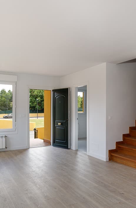 Entrada principal de una vivienda con suelo de madera clara, paredes blancas y escalera de madera hacia la planta superior. Puerta principal abierta con acceso directo al exterior, que muestra una zona ajardinada y luz natural abundante